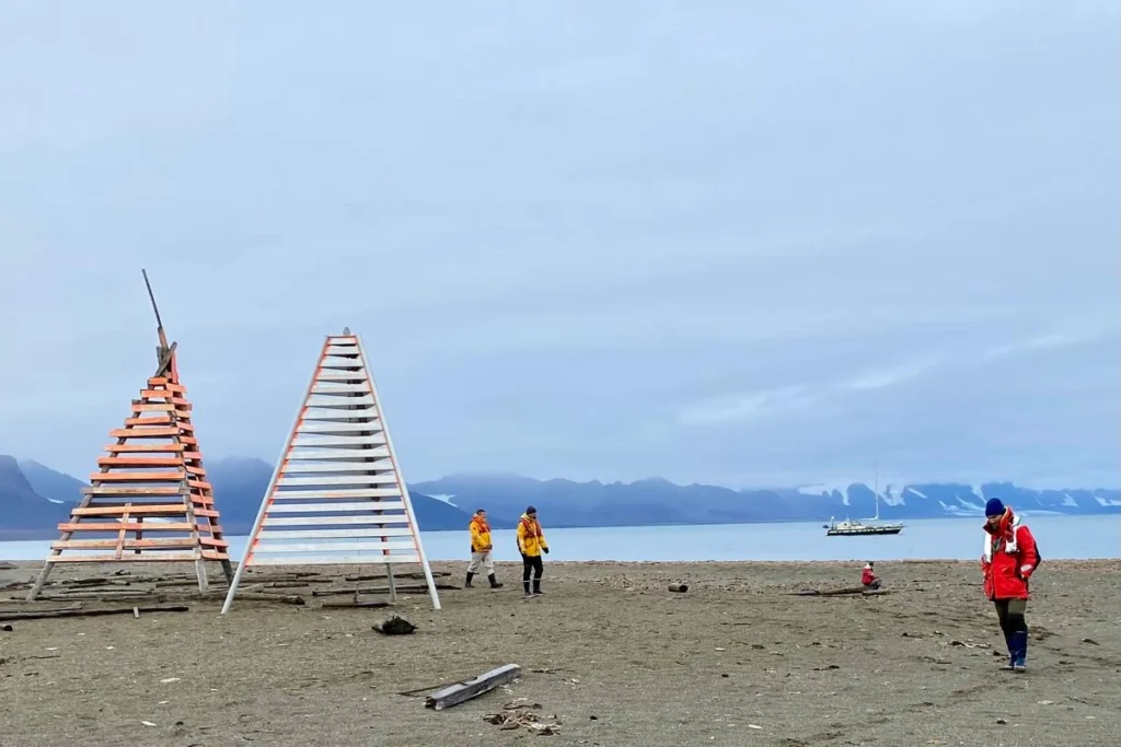 Załoga na plaży Poolepynten przy dwóch trójkątnych znakach nawigacyjnych, w tle jacht